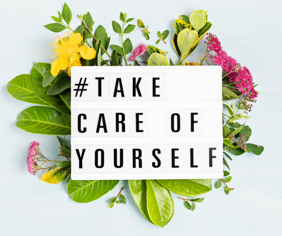 Letter board that reads "take care of yourself" on a bed of green leaves and flowers.