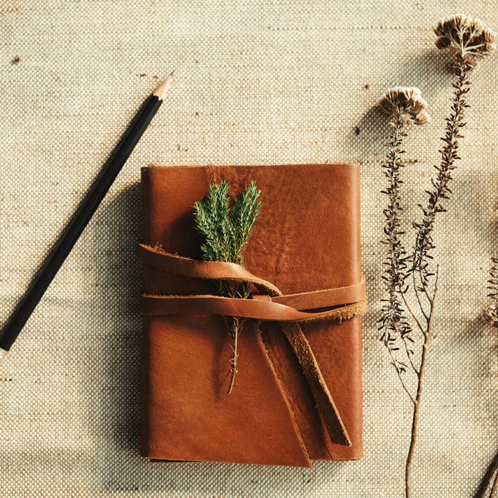A leather journal on a linen cloth with a pencil and some dried plant stems nearby.